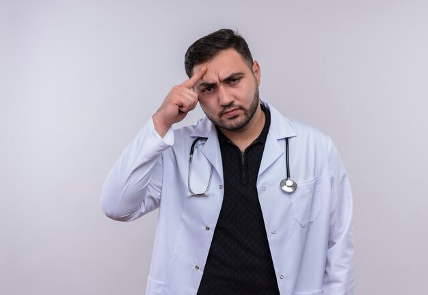 Joven médico varón barbudo con bata blanca con estetoscopio mirando a la cámara con cara seria disgustado