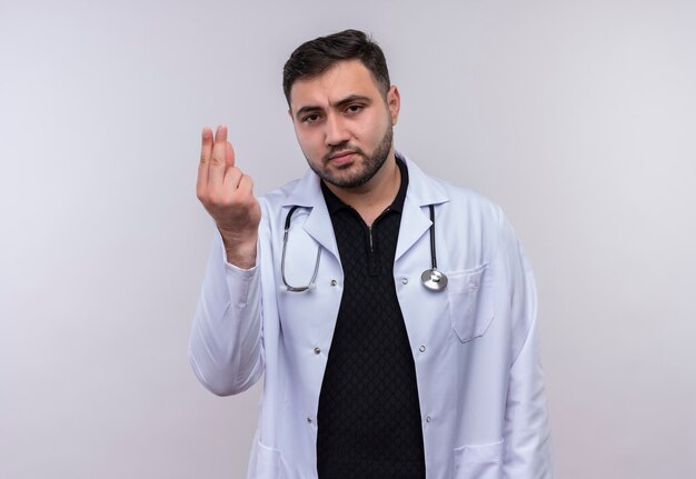 Joven médico varón barbudo con bata blanca con estetoscopio mirando a la cámara frotando los dedos gesto de dinero, pidiendo dinero