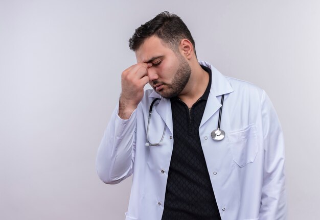 Joven médico varón barbudo con bata blanca con estetoscopio mirando cansado y aburrido tocando la nariz entre los ojos cerrados