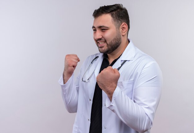 Joven médico varón barbudo con bata blanca con estetoscopio mirando a un lado posando como un boxeador con los puños cerrados sonriendo, concepto ganador