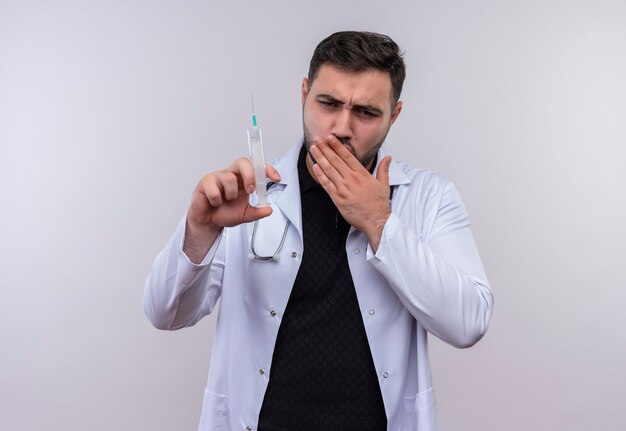 Joven médico varón barbudo con bata blanca con estetoscopio sosteniendo una jeringa mirando sorprendido cubriendo la boca con la mano