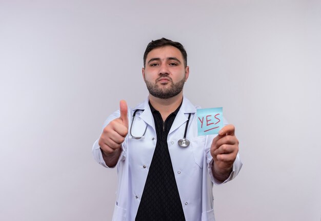 Joven médico varón barbudo con bata blanca con estetoscopio sosteniendo papel recordatorio con la palabra sí mostrando los pulgares para arriba