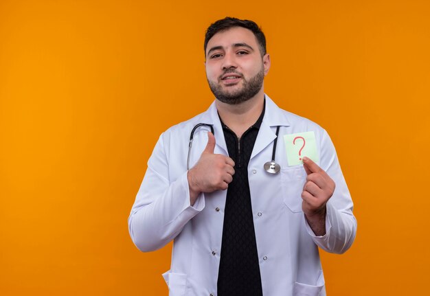 Joven médico varón barbudo con bata blanca con estetoscopio sosteniendo papel recordatorio con signo de interrogación sonriendo mostrando los pulgares para arriba