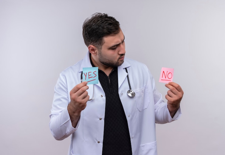 Foto gratuito joven médico varón barbudo con bata blanca con estetoscopio sosteniendo papeles recordatorios con palabras sí y no mirándolos intrigados