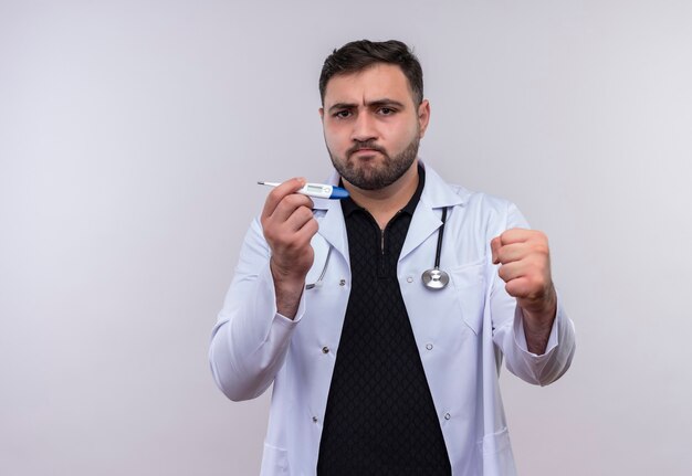 Joven médico varón barbudo con bata blanca con estetoscopio sosteniendo termómetro digital sonriendo confiado puño de apriete, concepto ganador