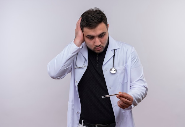 Foto gratuita joven médico varón barbudo con bata blanca con estetoscopio sosteniendo termómetro mirándolo muy emocional y preocupado