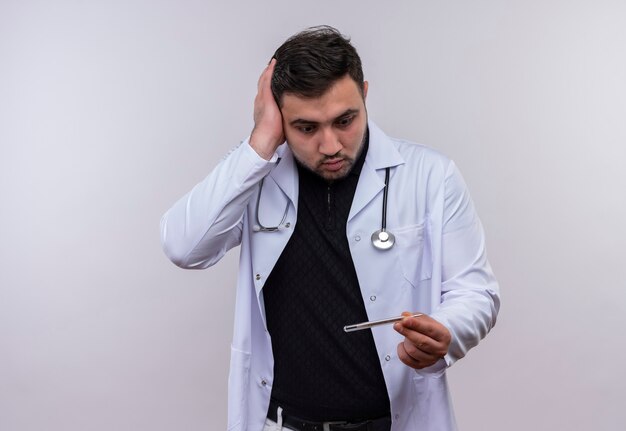Joven médico varón barbudo con bata blanca con estetoscopio sosteniendo termómetro mirándolo muy emocional y preocupado