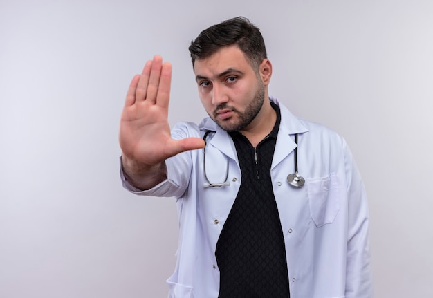 Joven médico varón barbudo vestido con bata blanca con estetoscopio con las manos abiertas haciendo señal de stop con cara seria