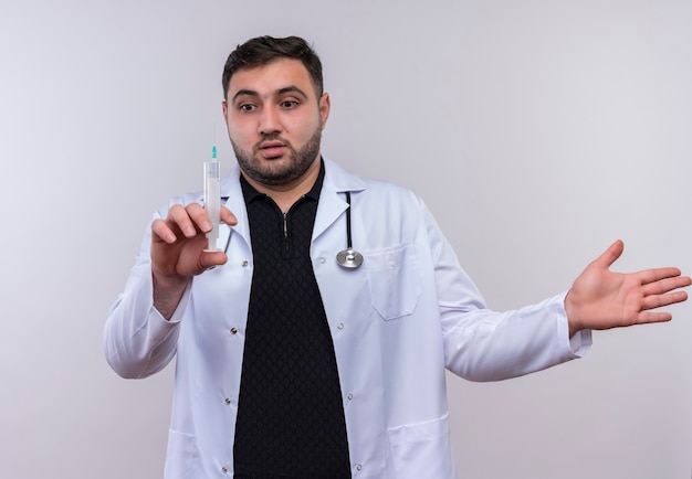 Joven médico varón barbudo vestido con bata blanca con estetoscopio sosteniendo una jeringa mirando incierto y confundido extendiendo el brazo hacia un lado