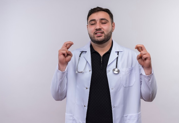 Joven médico varón barbudo vistiendo bata blanca con estetoscopio cerrando los ojos haciendo deseable con cruzar los dedos