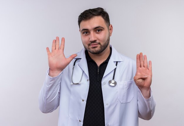 Joven médico varón barbudo vistiendo bata blanca con estetoscopio mostrando y apuntando hacia arriba con los dedos número nueve sonriendo confiado