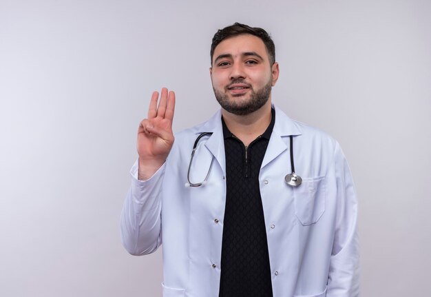 Joven médico varón barbudo vistiendo bata blanca con estetoscopio mostrando y apuntando hacia arriba con los dedos número tres sonriendo