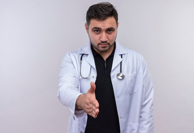 Joven médico varón barbudo vistiendo bata blanca con estetoscopio saludo con mirada de confianza ofreciendo mano