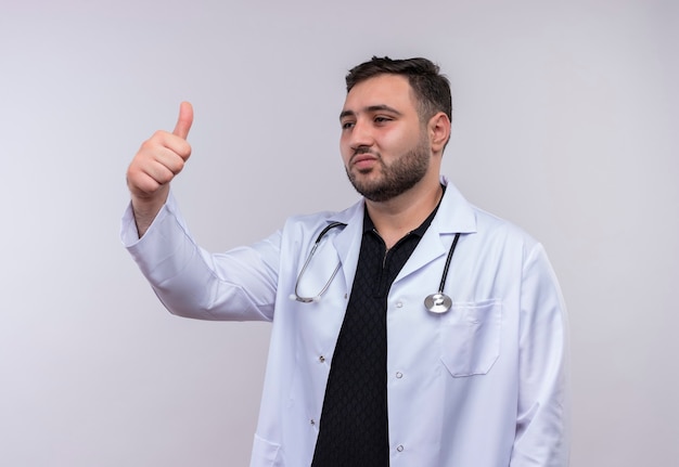 Joven médico varón barbudo vistiendo bata blanca con estetoscopio sonriendo mirando a un lado mostrando los pulgares para arriba