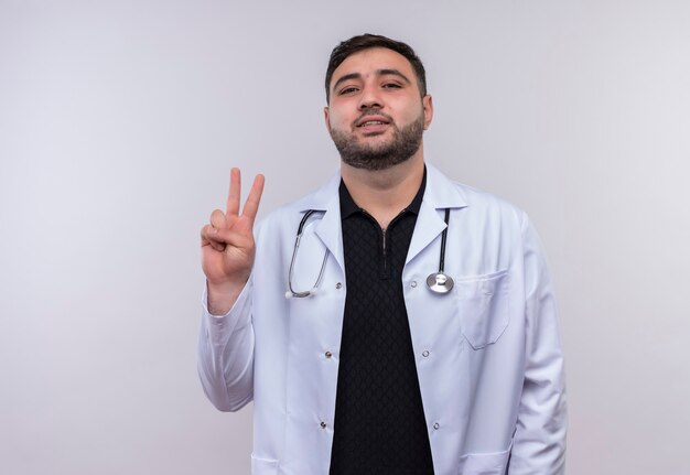 Joven médico varón barbudo vistiendo bata blanca con estetoscopio sonriendo mostrando el número dos o el signo de la victoria