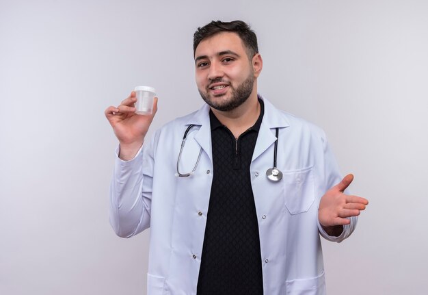 Joven médico varón barbudo vistiendo bata blanca con estetoscopio sosteniendo frasco de prueba sonriendo confiado