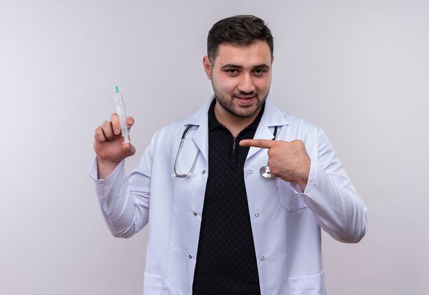 Joven médico varón barbudo vistiendo bata blanca con estetoscopio sosteniendo una jeringa apuntando con el dedo a ella sonriendo confiado