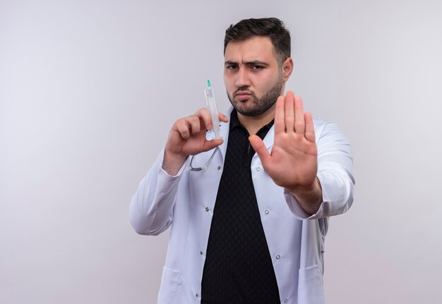 Joven médico varón barbudo vistiendo bata blanca con estetoscopio sosteniendo jeringa haciendo señal de stop con la mano con el ceño fruncido