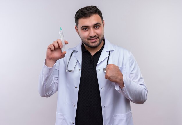 Joven médico varón barbudo vistiendo bata blanca con estetoscopio sosteniendo una jeringa sonriendo confiado apretando el puño, concepto ganador