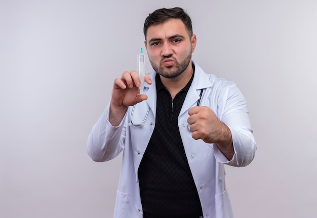 Joven médico varón barbudo vistiendo bata blanca con estetoscopio sosteniendo una jeringa sonriendo confiado apretando el puño, concepto ganador