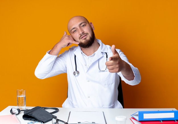 Joven médico varón calvo con bata médica y un estetoscopio sentado en el escritorio con herramientas médicas que muestran gestos de llamadas telefónicas y puntos a la cámara en naranja