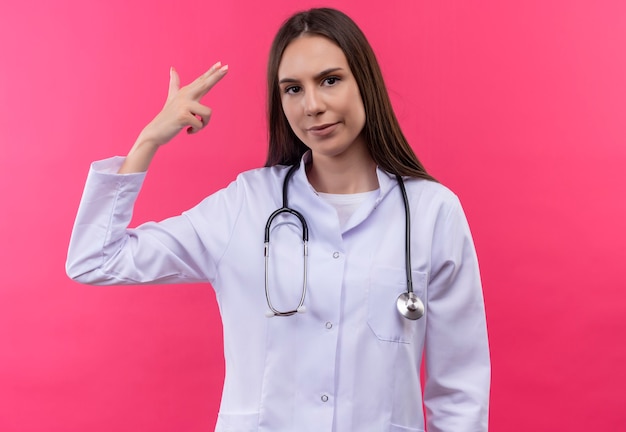 Joven médico vestida con estetoscopio bata médica mostrando gesto de pistola en pared rosa aislada