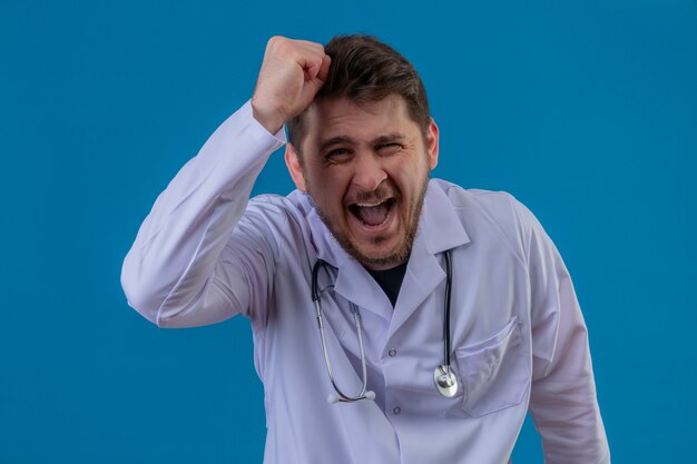 Joven médico vistiendo bata blanca y estetoscopio enojado y enojado levantando el puño frustrado y furioso mientras gritaba con ira sobre fondo azul aislado