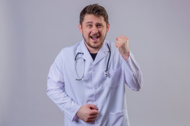 Joven médico vistiendo bata blanca y estetoscopio de pie con una gran sonrisa en la cara levantando el puño, concepto ganador