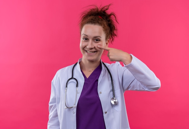 Joven médico vistiendo bata blanca con estetoscopio sonriendo con cara feliz apuntando con el dedo a la mejilla de pie sobre la pared rosa