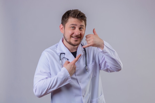 Joven médico vistiendo bata blanca y estetoscopio sonriendo haciendo hablar por el gesto del teléfono, llámame señal y señalando