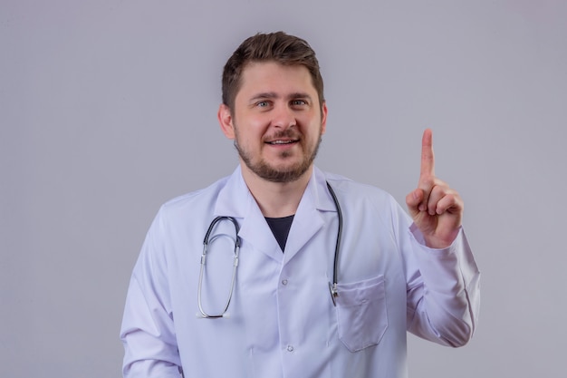 Joven médico vistiendo bata blanca y un estetoscopio con una sonrisa en la cara y apuntando con el dedo hacia arriba