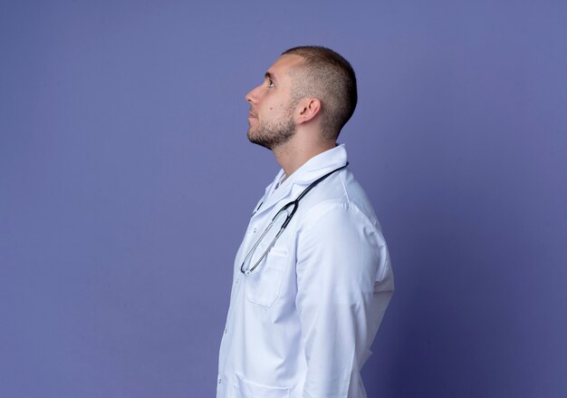 Joven médico vistiendo una bata médica y un estetoscopio alrededor de su cuello de pie en la vista de perfil y mirando hacia arriba aislado sobre fondo púrpura con espacio de copia