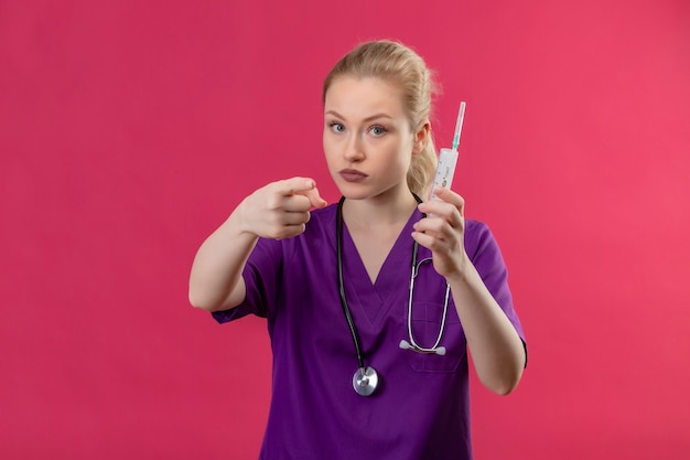 Joven médico vistiendo una bata médica púrpura y un estetoscopio con jeringa le muestra un gesto en la pared rosa aislada