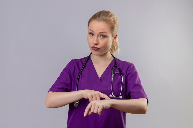 Joven médico vistiendo una bata médica púrpura y un estetoscopio muestra el gesto del reloj en la pared blanca aislada