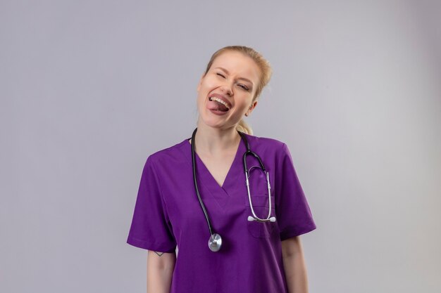 Joven médico vistiendo una bata médica púrpura y un estetoscopio muestra la lengua en la pared blanca aislada