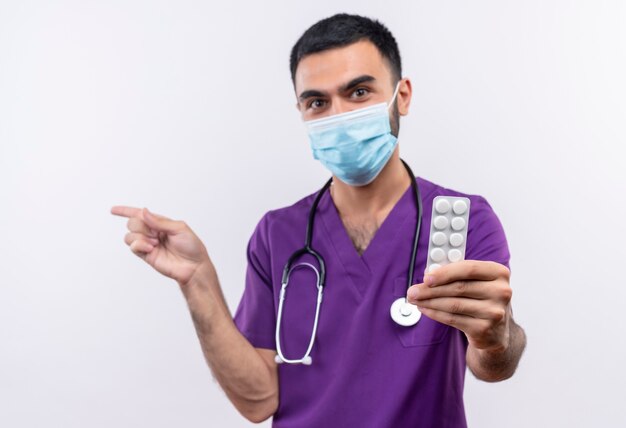 Joven médico vistiendo ropa de cirujano púrpura y estetoscopio máscara médica sosteniendo píldoras a la cámara apunta al lado de la pared blanca aislada