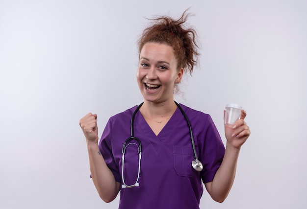 Joven médico vistiendo uniforme médico con estetoscopio sosteniendo el frasco de prueba feliz y salió apretando el puño parado sobre la pared blanca