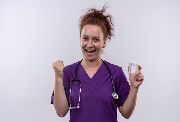 Joven médico vistiendo uniforme médico con estetoscopio sosteniendo el frasco de prueba feliz y salió apretando el puño parado sobre la pared blanca