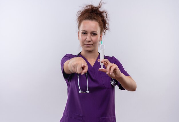 Joven médico vistiendo uniforme médico con estetoscopio sosteniendo una jeringa apuntando con el dedo mirando confiado de pie sobre la pared blanca