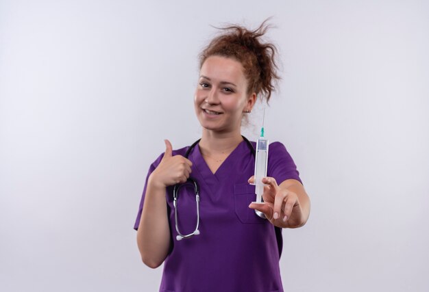 Joven médico vistiendo uniforme médico con estetoscopio sosteniendo una jeringa sonriendo y apuntando hacia atrás de pie sobre la pared blanca