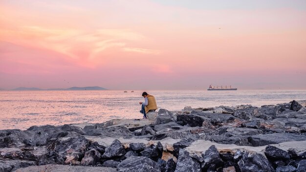 Joven meditando y leyendo en la orilla de una playa un barco pasando al atardecer