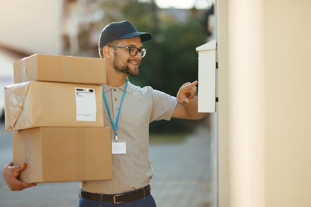 Joven mensajero feliz revisando el nombre en el buzón del cliente mientras entrega paquetes en el distrito residencial