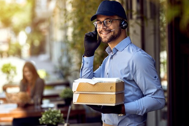 Joven mensajero feliz usando auriculares mientras entrega comida de un restaurante