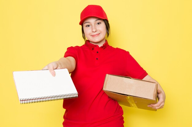 joven mensajero en polo rojo gorra roja con paquete cuaderno sonriendo en amarillo