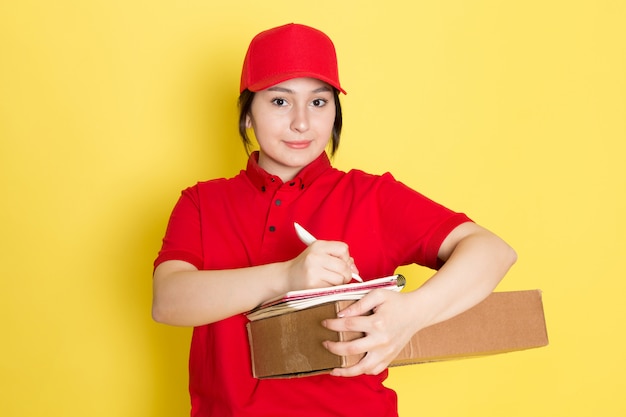 joven mensajero en polo rojo gorra roja con paquete escribiendo notas sonriendo en amarillo