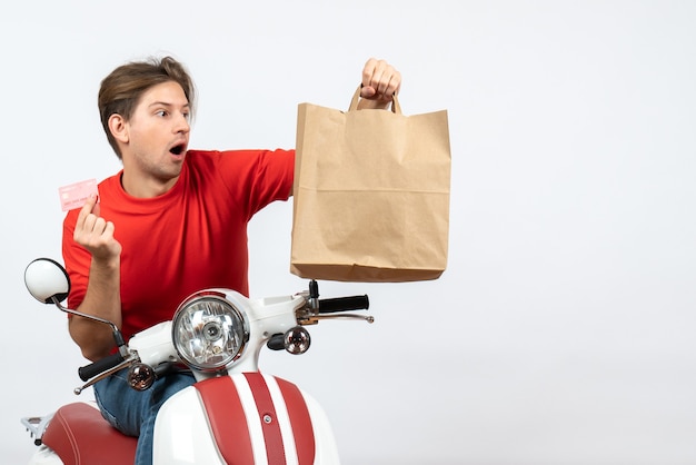 Joven mensajero sorprendido en uniforme rojo sentado en scooter sosteniendo una bolsa de papel en la pared amarilla