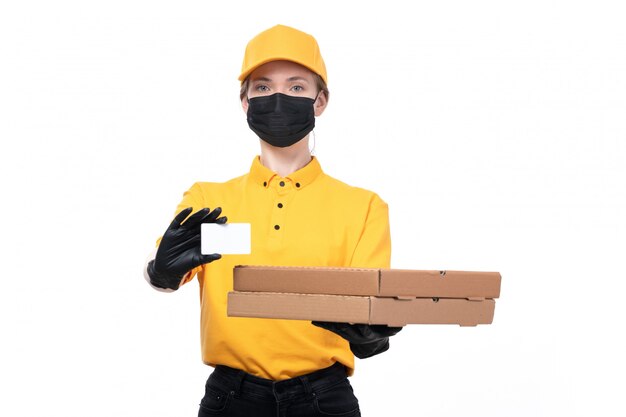 Una joven mensajero de vista frontal en uniforme amarillo guantes negros y máscara negra holdign tarjetas blancas y cajas de alimentos