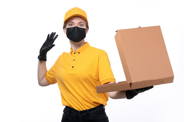 Una joven mensajero de vista frontal en uniforme amarillo guantes negros y máscara negra sosteniendo una caja de pizza vacía