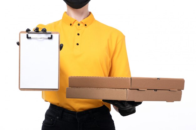 Una joven mensajero de vista frontal en uniforme amarillo guantes negros y máscara negra sosteniendo cajas de pizza y bloc de notas