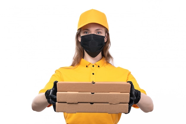 Una joven mensajero de vista frontal en uniforme amarillo guantes negros y máscara negra sosteniendo cajas de pizza